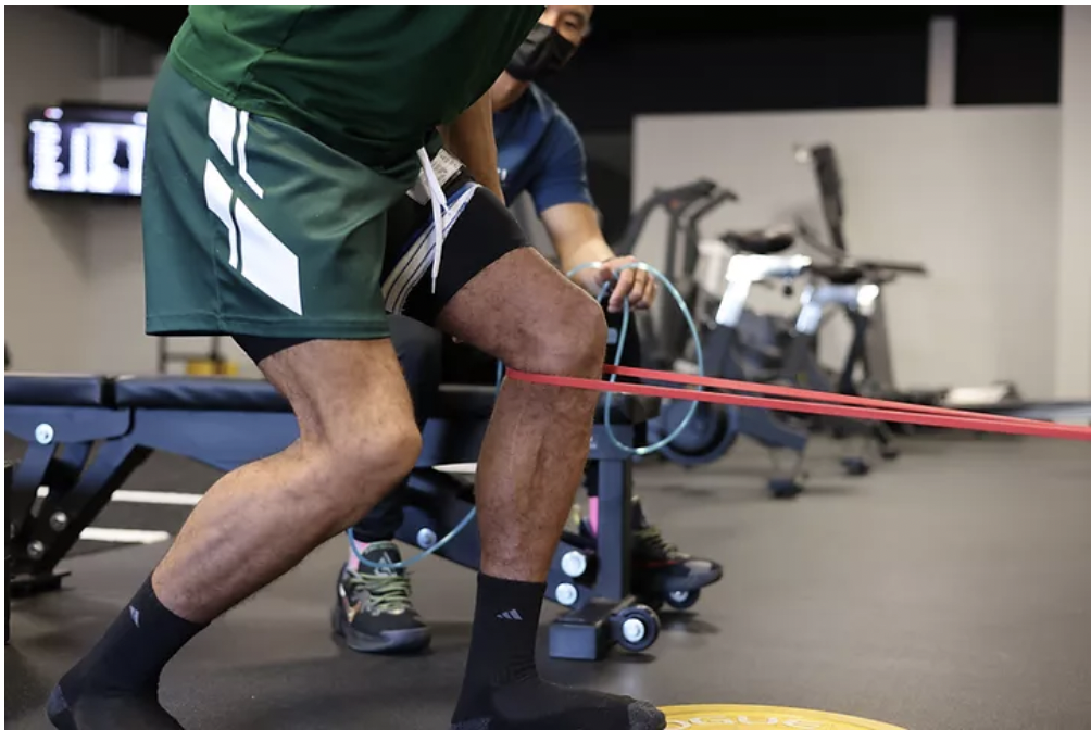 Weak knee joints exercise with a resistance band around the knee as a physical therapist coaches proper knee alignment in a clinic gym.